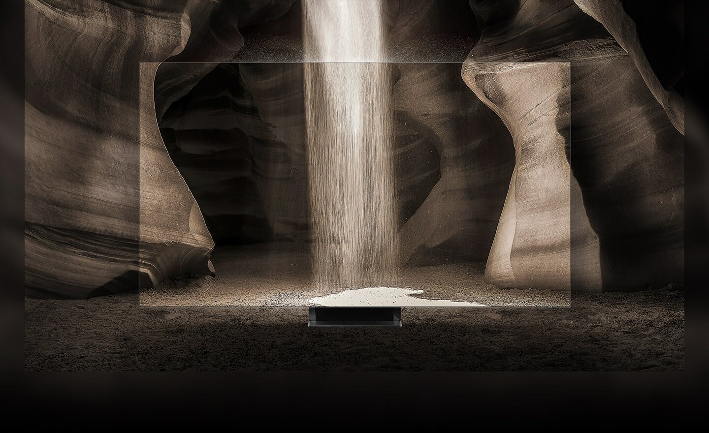 Telewizor QLED ustawiony w jaskini, z góry sypie się piasek. Krawędzie ekranu są tak wąskie, że wydaje się, jakby telewizora tam nie było. Telewizor QLED ustawiony w jaskini, z góry sypie się piasek. Krawędzie ekranu są tak wąskie, że wydaje się, jakby telewizora tam nie było.