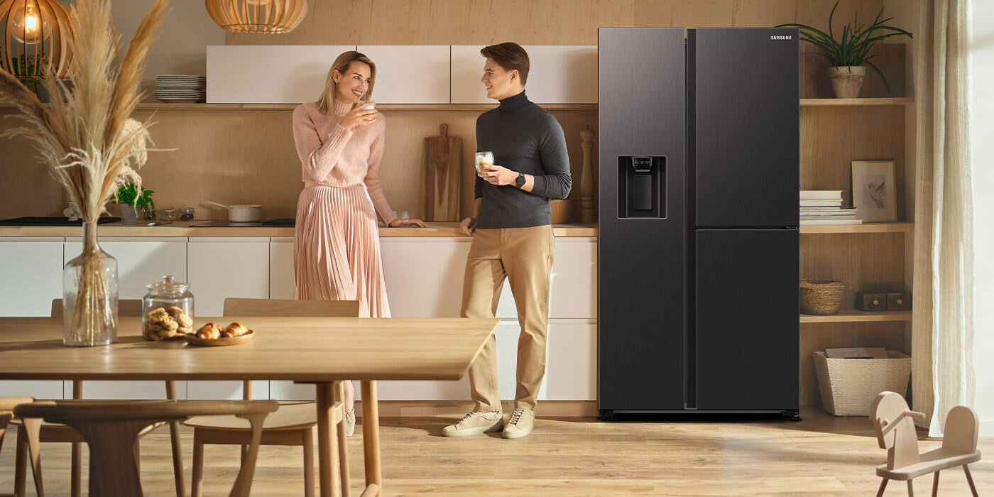 Ilustracja przedstawia kuchnię, w której większość elementów wyposażenia wykonana jest z jasnego drewna. W pomieszczeniu stoi lodówka Samsung w kolorze grafitowej stali, obok której znajdują się jej użytkownicy. Ilustracja przedstawia kuchnię, w której większość elementów wyposażenia wykonana jest z jasnego drewna. W pomieszczeniu stoi lodówka Samsung w kolorze grafitowej stali, obok której znajdują się jej użytkownicy.