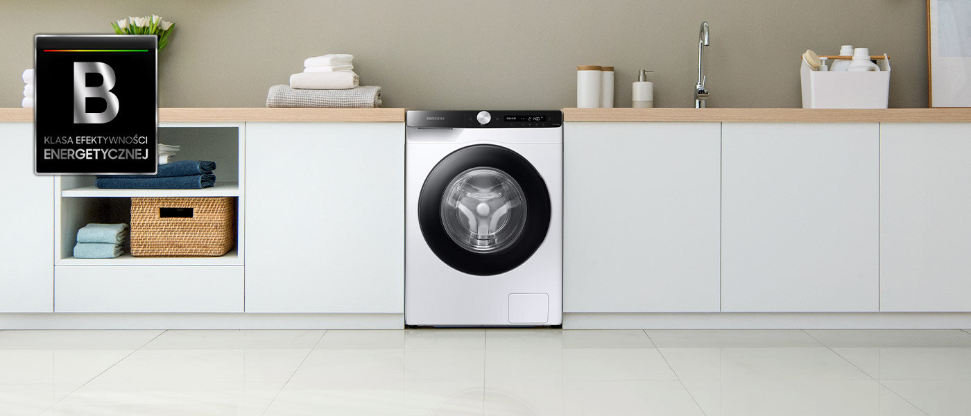 Biała pralka z czarnymi drzwiami stojąca w przestronnym pomieszczeniu, zdjęciu towarzyszy graficzny symbol klasy energetycznej B Biała pralka z czarnymi drzwiami stojąca w przestronnym pomieszczeniu, zdjęciu towarzyszy graficzny symbol klasy energetycznej B