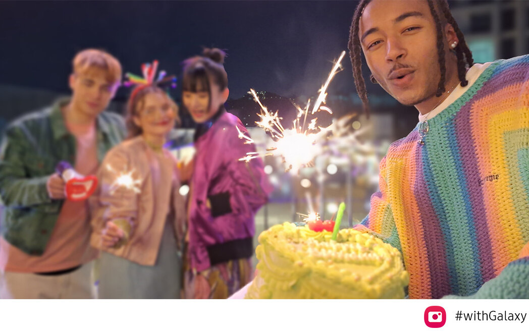 phpvcvbvy sm-a546-04-video Trzech przyjaciół znajduje się na szczycie mostu w nocy, świętując ze sztucznymi ogniami. Gdy w kadr wchodzi kolejny przyjaciel z prawej strony, trzymający tort, funkcja automatycznego kadrowania automatycznie dostosowuje kadr, aby objąć ich wszystkich, a także ustawić ostrość na przyjaciela trzymającego tort, który stoi bliżej w kadrze. Widoczna jest ikona aparatu i tekst #WithGalaxy.