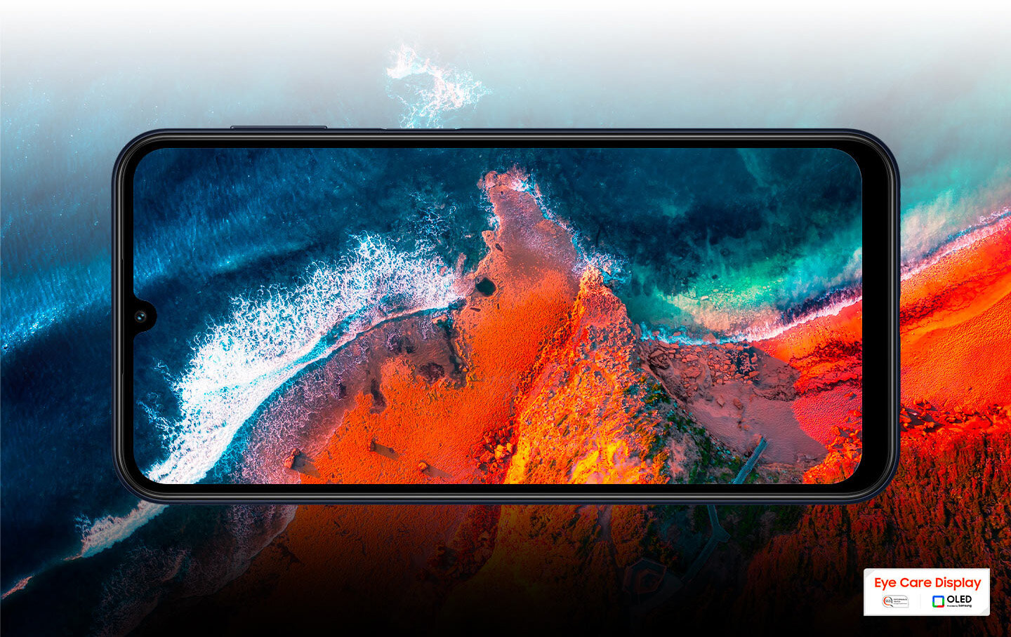 sm-m346-02screen1 Galaxy M34 5G pokazany z lotu ptaka nad plażą i piaskiem. Krajobraz w tle kontynuuje się na wyświetlaczu, przedzielony jedynie ramką urządzenia.