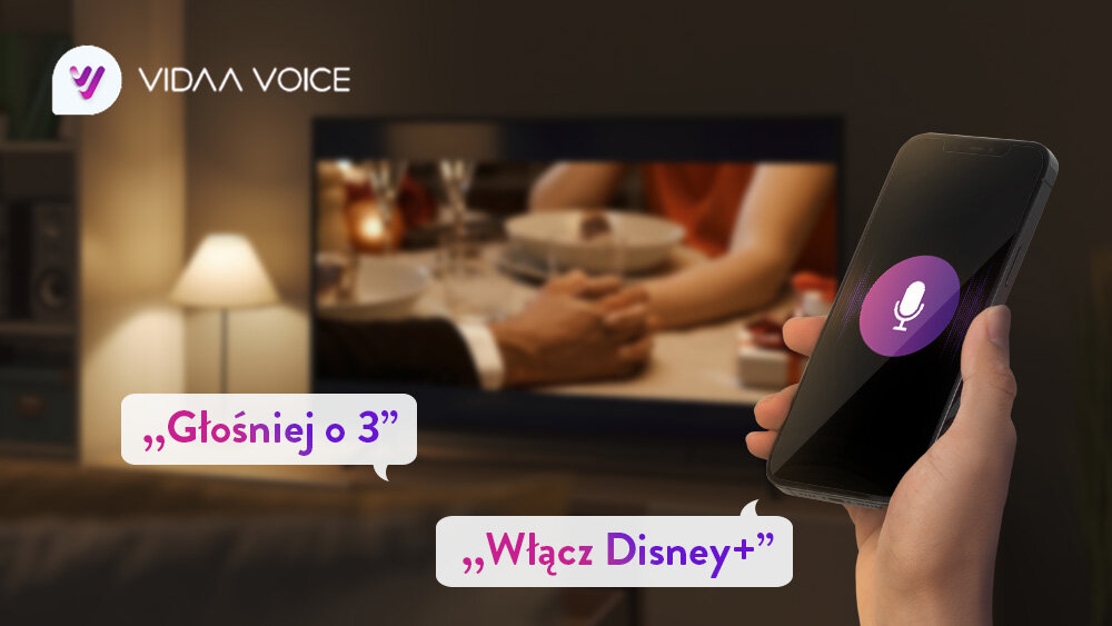 Telewizor JVC LT-65VD3500 - Szybkie sterowanie głosowe, telewizor stojący w salonie na stoliku, na pierwszym planie jest ręka z telefonem przedstawiający sterowanie głosowe